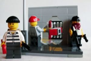 Lego minifigures staged as a bank robbery: a masked figure in striped prison clothes stands in front of an open vault while two others pass a red case between them, one of them armed.
