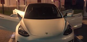 A white Tesla Model 3 at a charging stall at night with both front doors and the front trunk open.