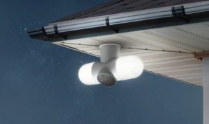 A white security camera with twin floodlights mounted under the eaves of a house against a night sky.