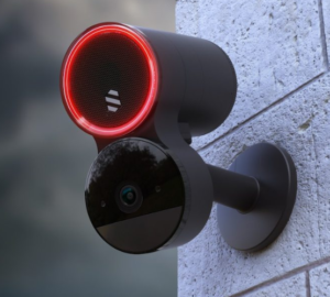 A wall-mounted outdoor security camera with a glowing red ring light around its upper sensor housing, fixed to grey brickwork.