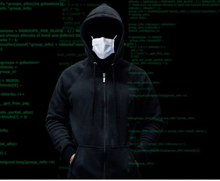 A figure in a black hooded sweatshirt and surgical mask standing in front of a screen of green source code, face not visible.