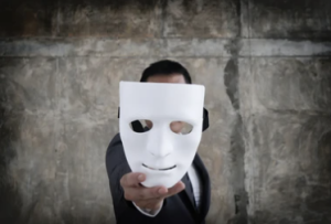 A man in a suit stands against a stone wall holding a blank white mask out toward the camera, his own face hidden behind it.
