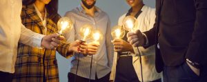 A circle of colleagues each holding a lit incandescent bulb, bringing them together at the centre of the group.